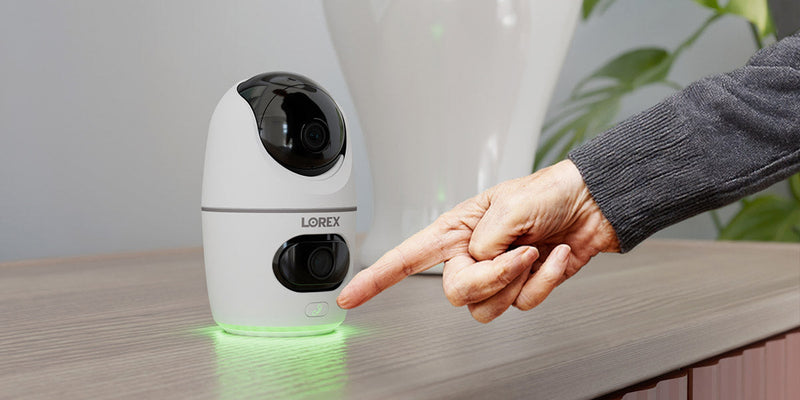 An elderly hand presses the call button on a Lorex 2K Dual Lens Indoor Security Camera, which glows green, signaling activation for emergency communication.