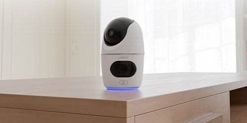 Lorex 2K indoor dual-lens pan-tilt security camera placed on a wooden table in a well-lit room, featuring a sleek white design with black camera lenses and a glowing blue light at the base.