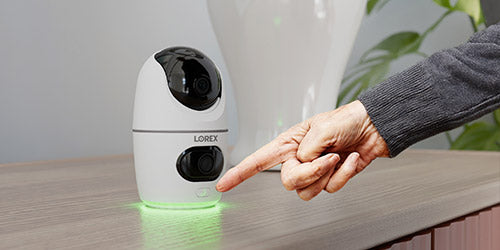 An elderly hand presses the call button on a Lorex 2K Dual Lens Indoor Security Camera, which glows green, signaling activation for emergency communication.