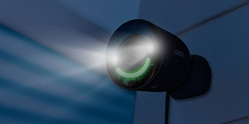 A black 4K outdoor security camera with a spotlight and green smart security lighting, mounted on a wall. The camera emits a bright white light, designed for enhanced visibility and nighttime security, with the brand logo "Lorex" visible on its side.